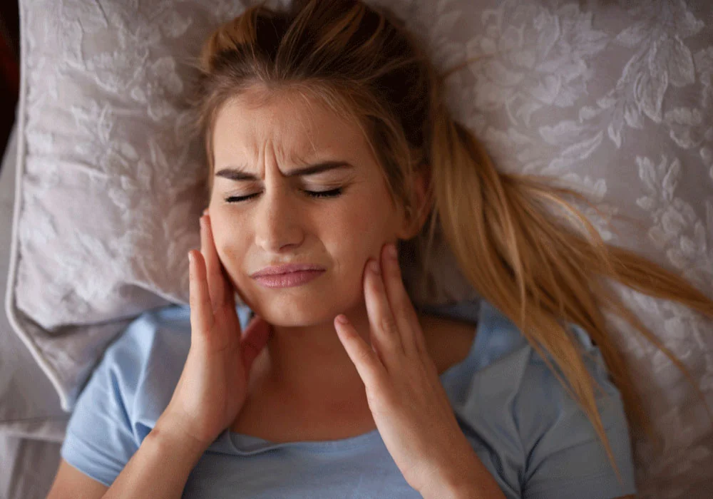 A woman lying on a bed with a patterned blanket and pillow, wearing a light blue shirt. She has long, tied-back blonde hair, and their hands are placed on their cheek.